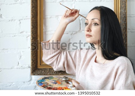 Young woman with long black hair holds a brush and a palette with multi-colored acrylic paints on the background of an empty vintage frame, selective focus