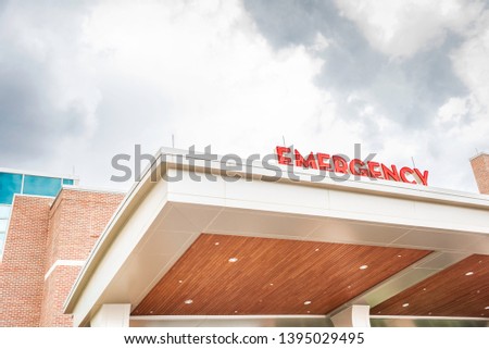 A prominently visible 3-D lighted all caps "emergency" signage in red atop the canopy of a hospital entrance in horizontal image format.