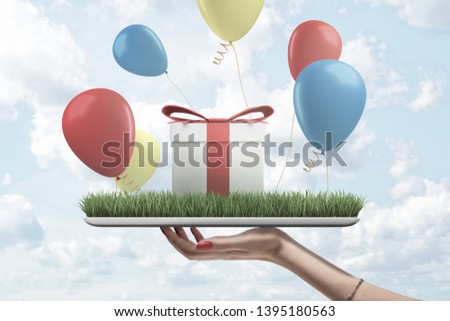 Side view of woman's hand holding digital tablet with green grass on screen, gift box standing on top and colorful balloons flying around. Giving presents. Having fun. Festivals and celebrations.