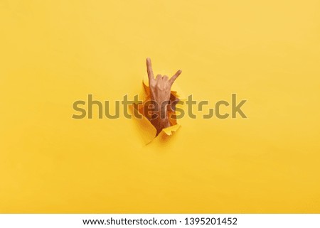 Unrecognizable man makes rock n roll gesture through ripped hole in yellow paper. Male demonstrates horn sign with hand stretched in gap slot of paper. Body language concept. Colored background Royalty-Free Stock Photo #1395201452
