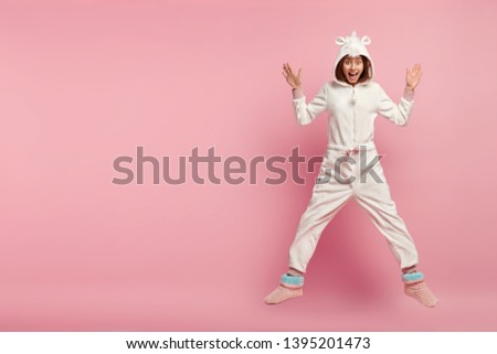 Indoor shot of overjoyed happy girl in comfortable domestic kigurumi costume, jumps over pink background, has hoody on head, enjoys weekend at cosy home, isolated over rosy wall with empty space Royalty-Free Stock Photo #1395201473