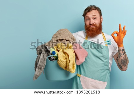 Satisfied bearded red haired man makes okay gesture, shows approval sign, says everything is under control, busy with daily routines, holds basin with dirty laundry, isolated on blue background