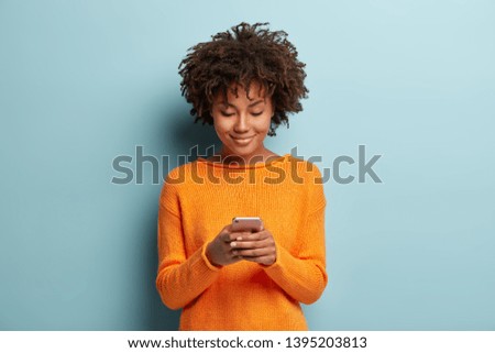 Horizontal shot of beautiful satisfied female teenager focused in smartphone device, chats online with friends, checks notification received on email, being always in touch, wears casual orange jumper Royalty-Free Stock Photo #1395203813