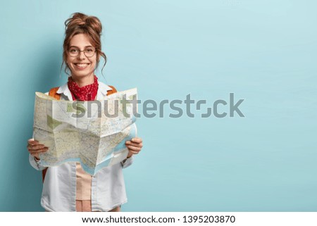 Positive smiling young female tourist searches for inspiring places, holds paper map, finds new sightseeing for discovering, wears round spectacles, red bandana, isolated on blue studio wall