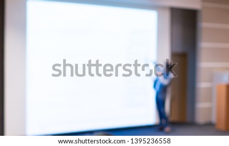 Blurred business presentation with keynote speaker near blank white screen. Seminar concept background with executive leading workshop in conference hall. Presenter in lecture to audience.