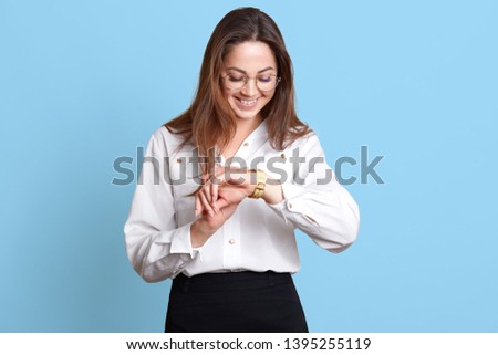 Studio shot of happy smiling beautiful brunette young woman in formal clothes standing and checking time, being in hurry, has important meeting, isolated on blue background. Business concept.