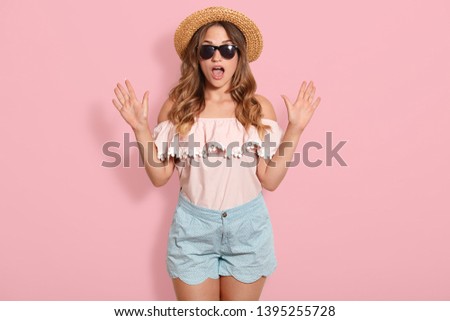 Image of young woman wearing pink blouse, blue short, sunglasses and summer straw hat standing with open mouth in shock, keeps hands up, has astonished facial expression. Human emotions concept.