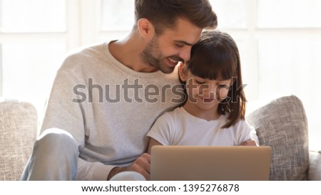 Loving father teaching little daughter to use laptop at home, sitting together on couch, smiling parent with adorable child playing computer game or making video call, spending free time together