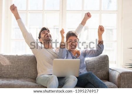 Happy young couple celebrate online victory, screaming with joy raising hands, looking at laptop screen together, wife and husband receive good news, achievement, new great offer opportunity, success Royalty-Free Stock Photo #1395276917