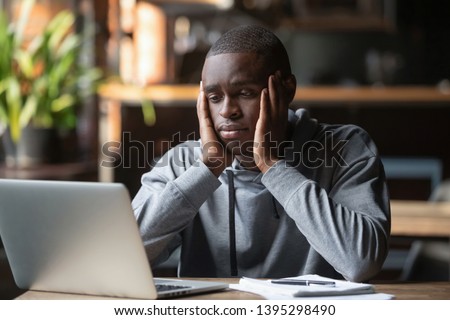 Tired African American man working on laptop in coffeehouse, holding head in hands, looking at screen, exhausted student taking break, watching webinar, working on research work or homework in cafe Royalty-Free Stock Photo #1395298490