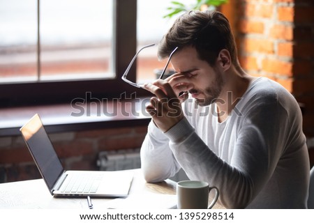 Upset man massaging nose bridge, taking off glasses, feeling eye strain after long work with laptop, sitting in cafe, tired male feeling discomfort after long wearing glasses, bad eye vision concept Royalty-Free Stock Photo #1395298514