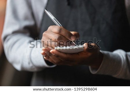 Close up African American waiter hands with notebook, taking customer order in cafe or restaurant, coffeehouse worker wearing black apron serving client, writing down, service concept Royalty-Free Stock Photo #1395298604