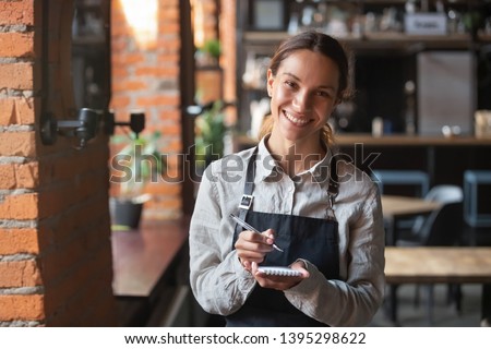 Head shot portrait of smiling waitress wearing black apron ready to take customer order, attractive woman with notebook and pen in hands looking at camera, standing in cozy coffeehouse, good service Royalty-Free Stock Photo #1395298622