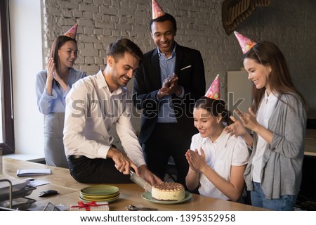 Happy birthday boy hold knife cutting cake feels gratitude for attention surprise. Diverse colleagues wear party hat glad to congratulate workmate best wishes friendly warm relations at work concept