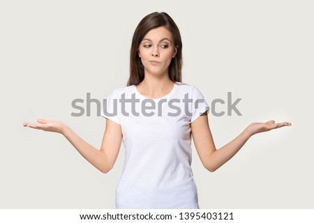 Young funny woman wearing white t-shirt stretched hands feels confused pose isolated on grey wall, girl imagining alternatives, weighs pros and cons, choosing make not easy difficult decision concept Royalty-Free Stock Photo #1395403121