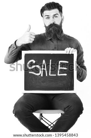 Bearded man, long beard. Brutal caucasian amused hipster with moustache holding sale inscription on blackboard, sitting on orange chair wearing blue shirt and vinous pants isolated on white background