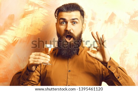 Bearded man, long beard. Brutal caucasian surprised hipster with moustache in brown shirt holding alcoholic red shot on colorful texture background