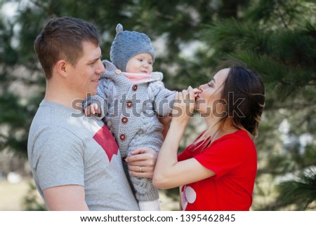 Beautiful woman and man with baby on walk. Mother in a red T-shirt kissing hand her baby in hand-knitted overall. Coniferous background.