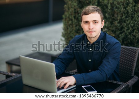 Businessman using laptop in cafe. Handsome man in dark shirn sit on the terrace. Background of green tree and blue sky