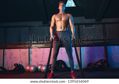 Caucasian bodybuilder practicing effective way to torch calories and enhance cardiovascular health. Full length photo. Battle rope training in a gym