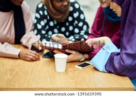 Photo of a happy muslim multiethnic girls students discussing book, sitting at table with take-away tea in colledge hall.