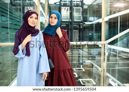 Portrait of two islamic young women dressed in muslim wear with hijabs on head looking at camera while standing against glass and steel loft interior