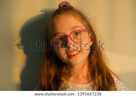 
baby girl with brown hair and glasses in a white t-shirt along the white wall