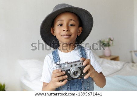 Art, technology, hobby and childhood concept. Handsome joyful dark skinned black schoolboy interested in photography, holding vintage film camera, smiling, wearing elegant round hat in bedroom