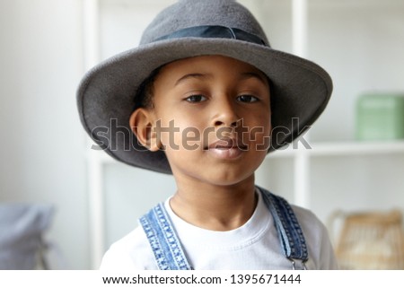 Style, beauty, children’s wear and fashion concept. Close up picture of adorable fashionable eight year old Afro American boy posing indoors wearing stylish round men's hat and denim jumpsuit