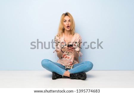 Young blonde woman sitting on the floor surprised and sending a message