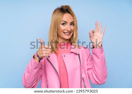 Young blonde woman with pink jacket over isolated blue background showing ok sign and thumb up gesture