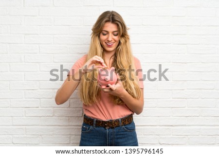Young blonde woman over white brick wall holding a big piggybank