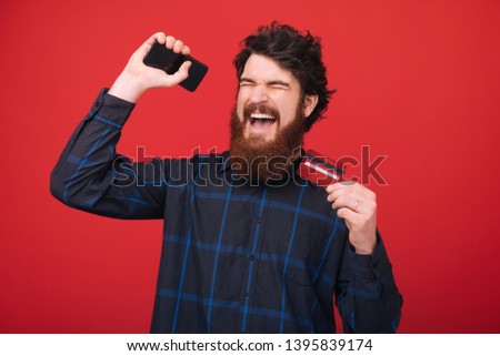Photo of bearded man, celebrating his first salary, with rised hands and closed eyes, red isolated background 