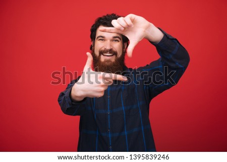 Photo of handome bearded man standing over red background and doing a photo frame with fingers, taking a photo 