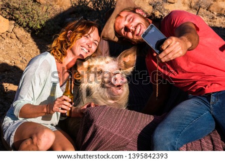 Happy alternative nature animal lover couple of cheerful people enjoy and have fun taking selfie picture with a funny pig in friendship - different lifestyle for millennials outdoor