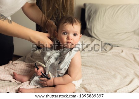 A young mother puts her baby in baby clothes. The baby obediently sits on the bed. Authentic lifestyle photos.