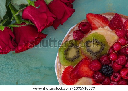 Flat-lay of red flowers roses and beautiful delicious sweet cake with berries on a green background. Greeting card of flowers roses and a cake with fruits. Copy space for text. Closeup or macro image.