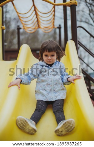 childhood: girl two years old brunette in a dress and jacket riding on a yellow children's slide on the playground