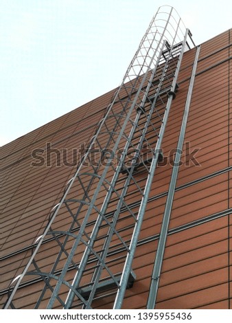 Stairway to the sky. Metal fire escape staircase from roof on a modular paneled wall of industrial or office building. Abstract modern architecture photo. Fire safety.