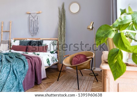 Stylish and luxury interior of bedroom with design furnitures, honey yellow armchair, gray macrame, mirror on the wall and elegant accessories. Beautiful bed sheets, blankets and pillows. Home decor.