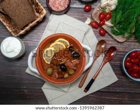 Russian traditional soup solyanka with meat and sausages, olives, tomatoes, lemons and mushrooms in a white bowl on a wooden table