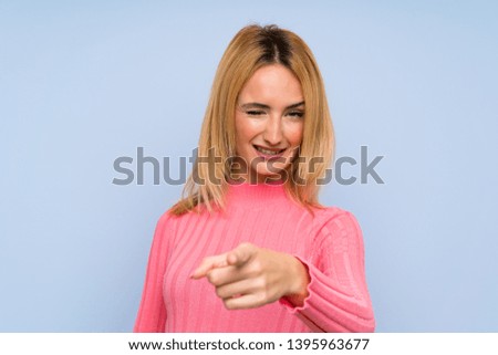 Young blonde woman with pink sweater over isolated blue background points finger at you