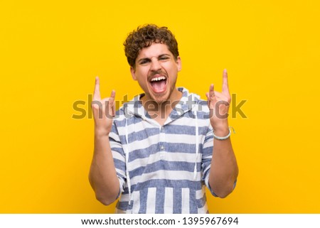 Blonde man over yellow wall making rock gesture