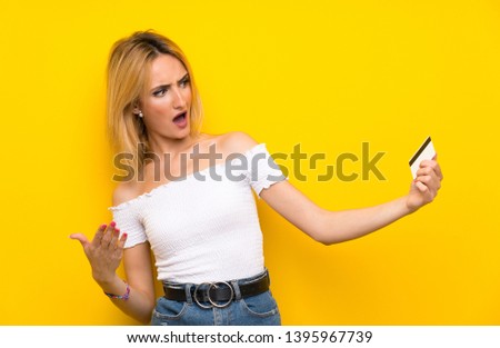 Young blonde woman over isolated yellow wall holding a credit card