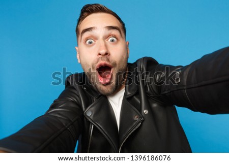 Close up selfie of stylish young unshaven man in black leather jacket white t-shirt looking camera isolated on blue background studio portrait. People sincere emotions concept. Mock up copy space