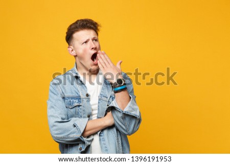 Boring tired young man in denim casual clothes looking aside, covering mouth with hand, yawning isolated on yellow orange background. People sincere emotions, lifestyle concept. Mock up copy space