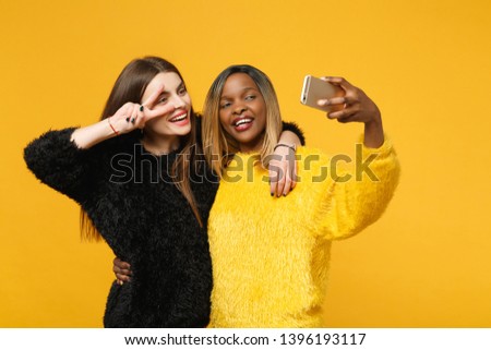 Young women friends european, african american in black yellow clothes hold in hand cellphone isolated on bright orange wall background, studio portrait. People lifestyle concept. Mock up copy space