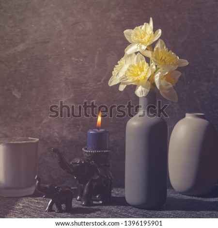 A bouquet of daffodils is in a tall gray bottle. Nearby are small figures of an elephant and a baby elephant. On the elephant is a burning blue candle. Behind is a vase. Gray stone background.