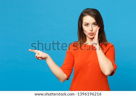 Beautiful young woman in red orange dress standing posing pointing hands fingers on workspace isolated over trendy blue wall background, studio portrait. People lifestyle concept. Mock up copy space