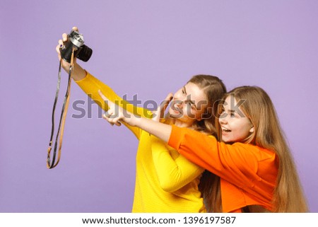 Two funny young blonde twins sisters girls in colorful clothes doing selfie shot on retro vintage photo camera isolated on violet blue background. People family lifestyle concept. Mock up copy space